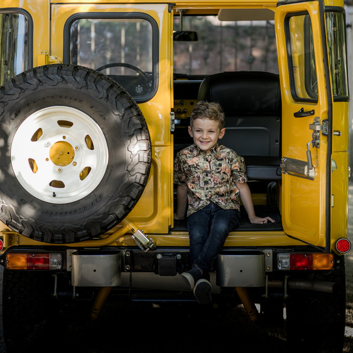 Smiling boy wears a Pladra Toddler Cubs Outdoor Aloha Shirt–Mountain Lions, sitting in back of yellow car with spare tire.