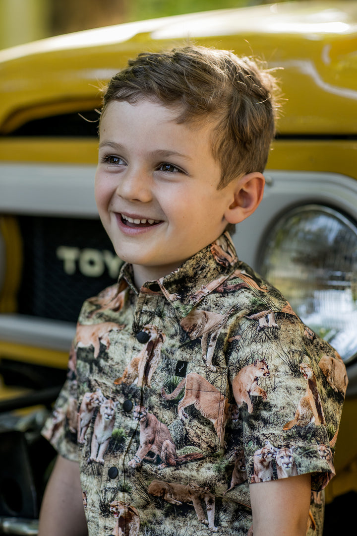 Smiling boy wears a Pladra Toddler Cubs Outdoor Aloha Shirt – Mountain Lions, standing by a yellow Toyota.