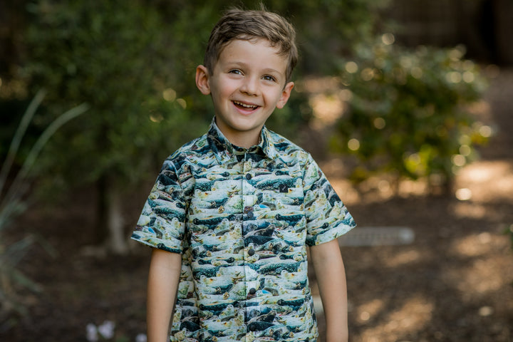 A young boy with light brown hair smiles while standing outdoors in a 100% cotton Toddler Cubs Outdoor Aloha Shirt–Seals, surrounded by greenery and soft, natural light.