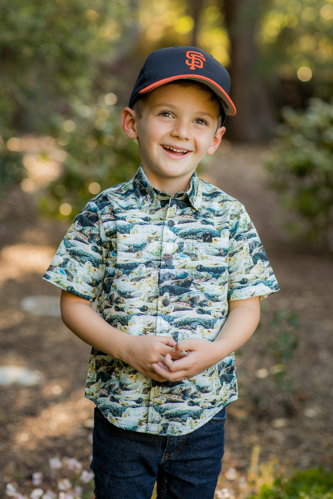 A young boy stands outdoors, smiling. He wears a black San Francisco Giants cap, dark jeans, and the Toddler Cubs Outdoor Aloha Shirt- Seals—a 100% cotton button-up featuring fish prints—set against blurred greenery and sunlight.