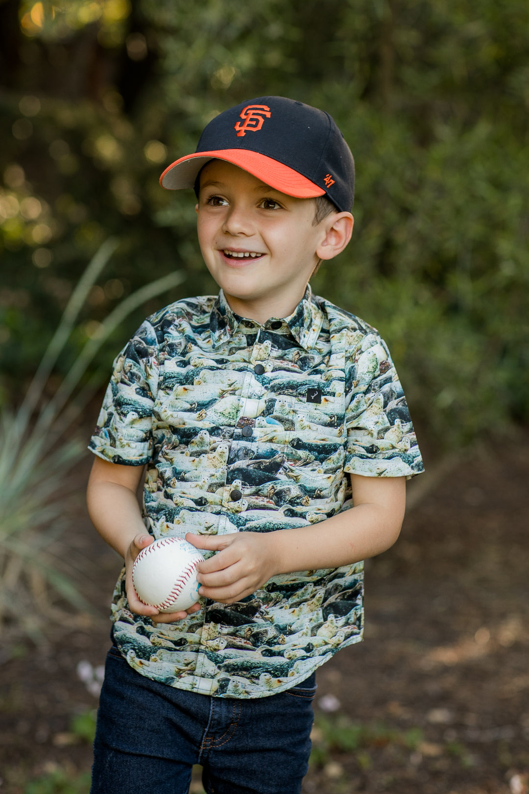 A young boy wearing a San Francisco Giants cap and the Toddler Cubs Outdoor Aloha Shirt- Seals smiles while holding a baseball, standing outdoors with greenery in the background.