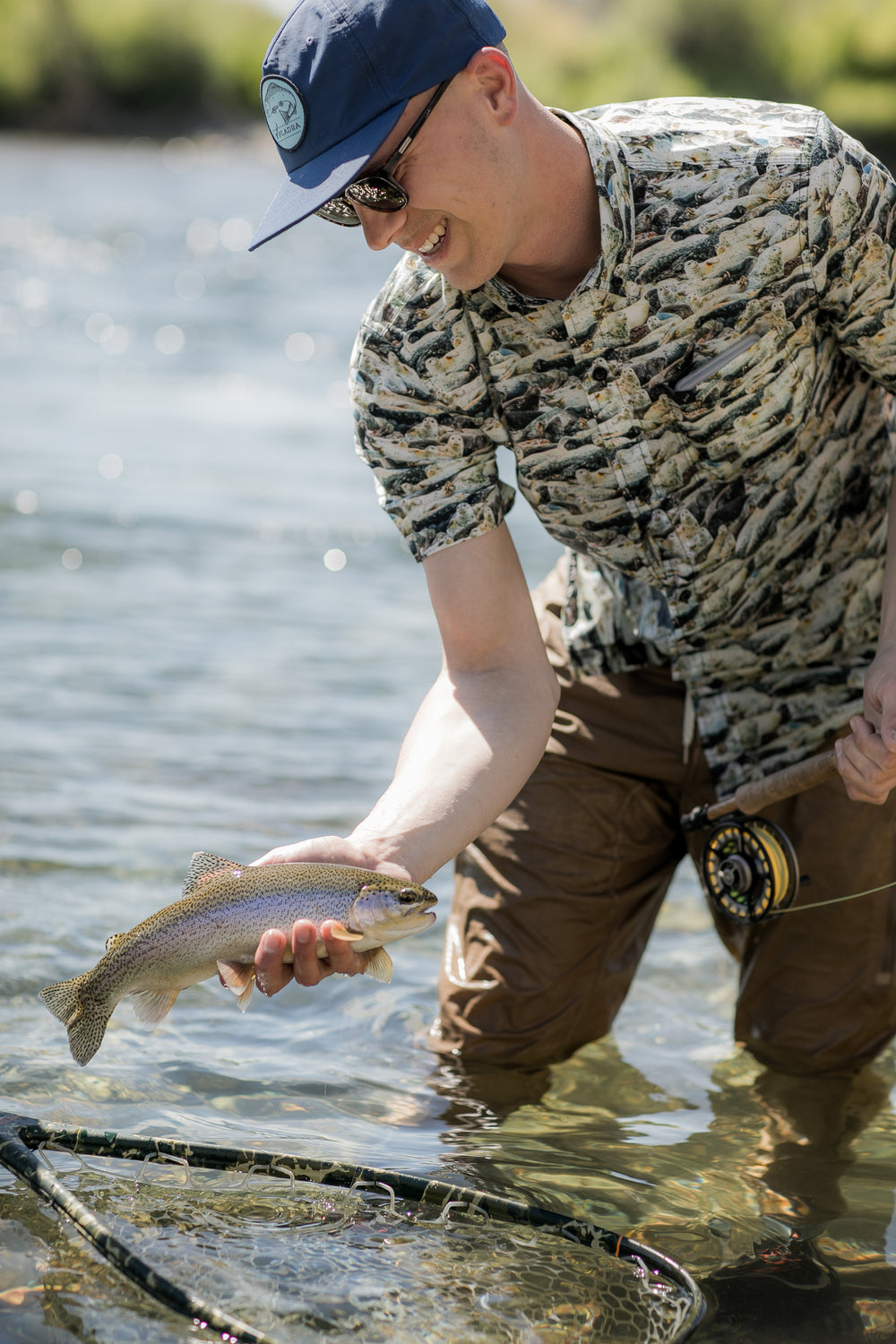 Man in sunglasses holds a trout riverside, wearing Pladra's Men's S/S Printed Outdoor Aloha Shirt - Seals and a blue cap.
