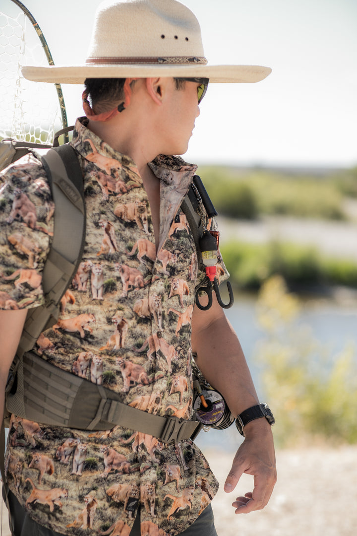 A person in a wide-brimmed hat, sunglasses, and backpack stands outdoors wearing the Men&