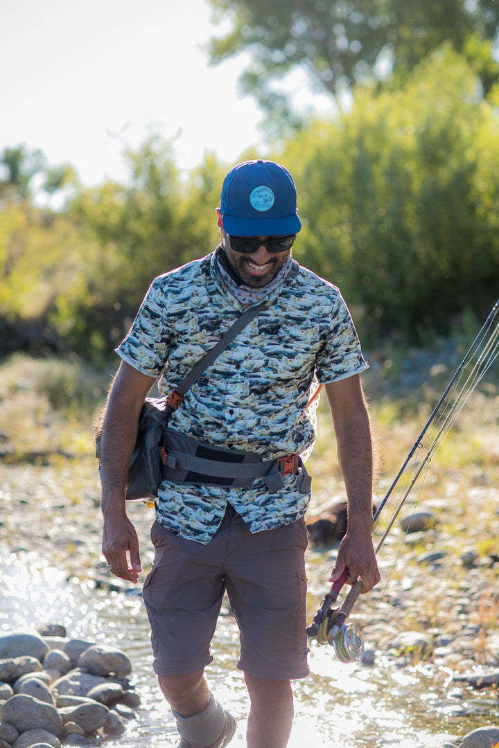 Wearing a Nylon Trout Hat in navy blue and sunglasses, a man smiles while fly fishing in a shallow, rocky stream on a sunny day. He carries quick-drying fishing gear and stands near the water’s edge.