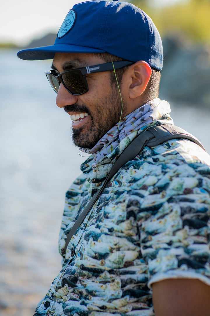 A man in sunglasses and a patterned shirt smiles by the water on a sunny day, wearing the Nylon Trout Hat in Navy Blue.