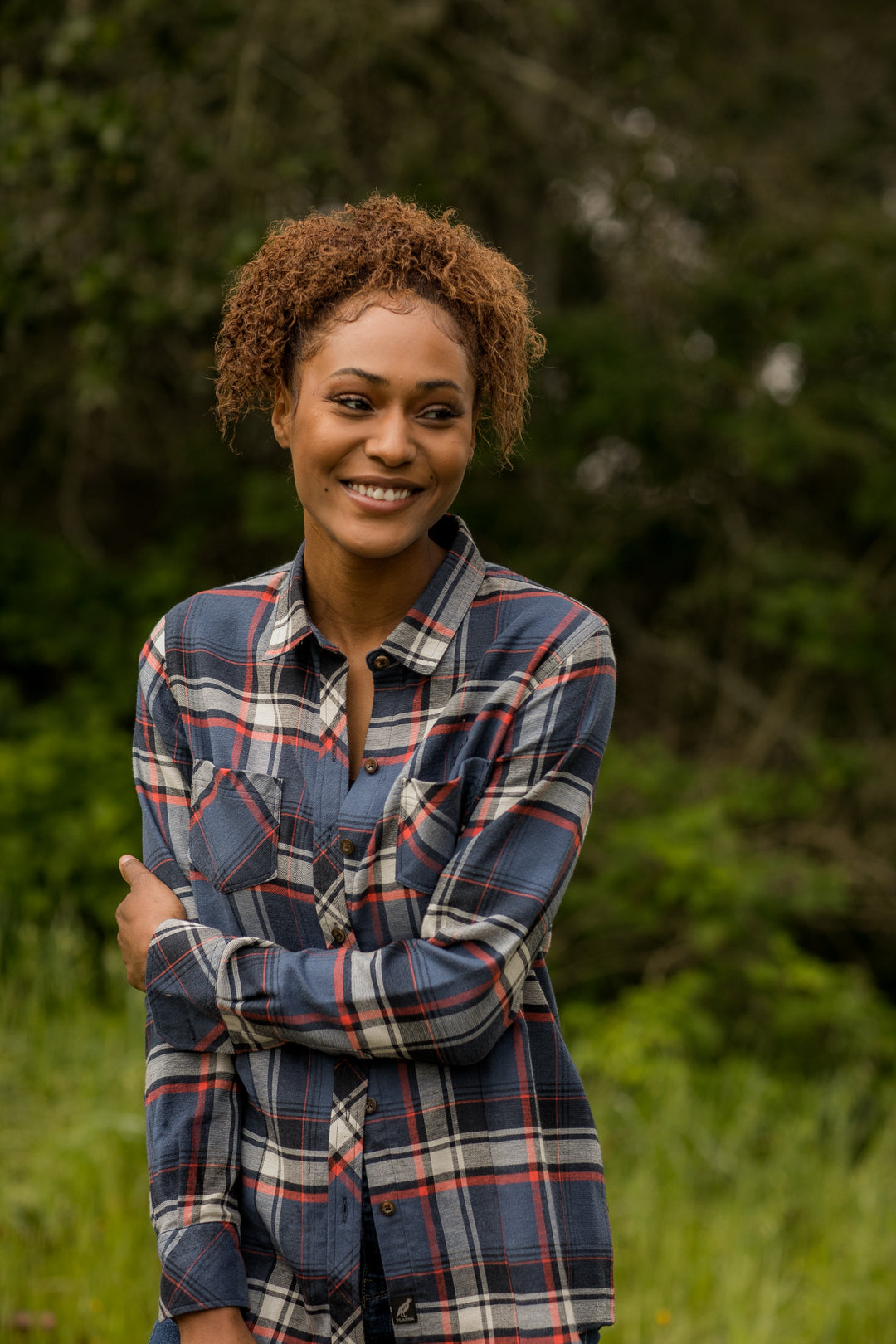 A woman with curly hair stands outside, smiling and looking to the side, wearing the Women&