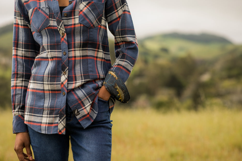 A woman stands in a grassy field wearing the Women's Every Day Double Weave Shirt in Sunset Blue, paired with jeans and a leather bracelet. She has one hand in her pocket; green hills and a cloudy sky blur in the background.