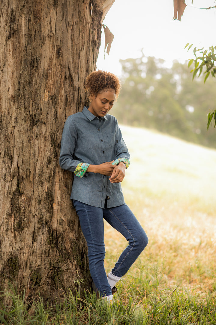 A woman with curly hair stands outdoors by a tree, wearing the Women&