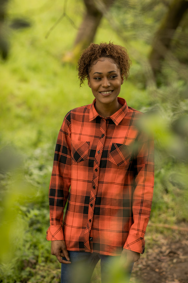 A woman with curly hair smiles outdoors among trees, wearing the Women&