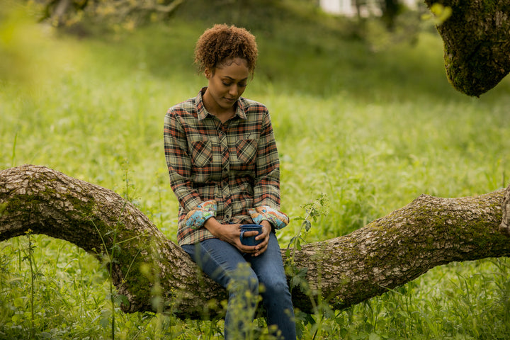 Woman sitting on a tree branch in a grassy field, holding a mug and looking down thoughtfully.