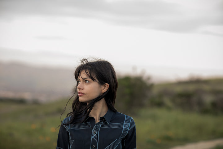A young woman with dark hair stands in a grassy field under cloudy skies, wearing the Women&