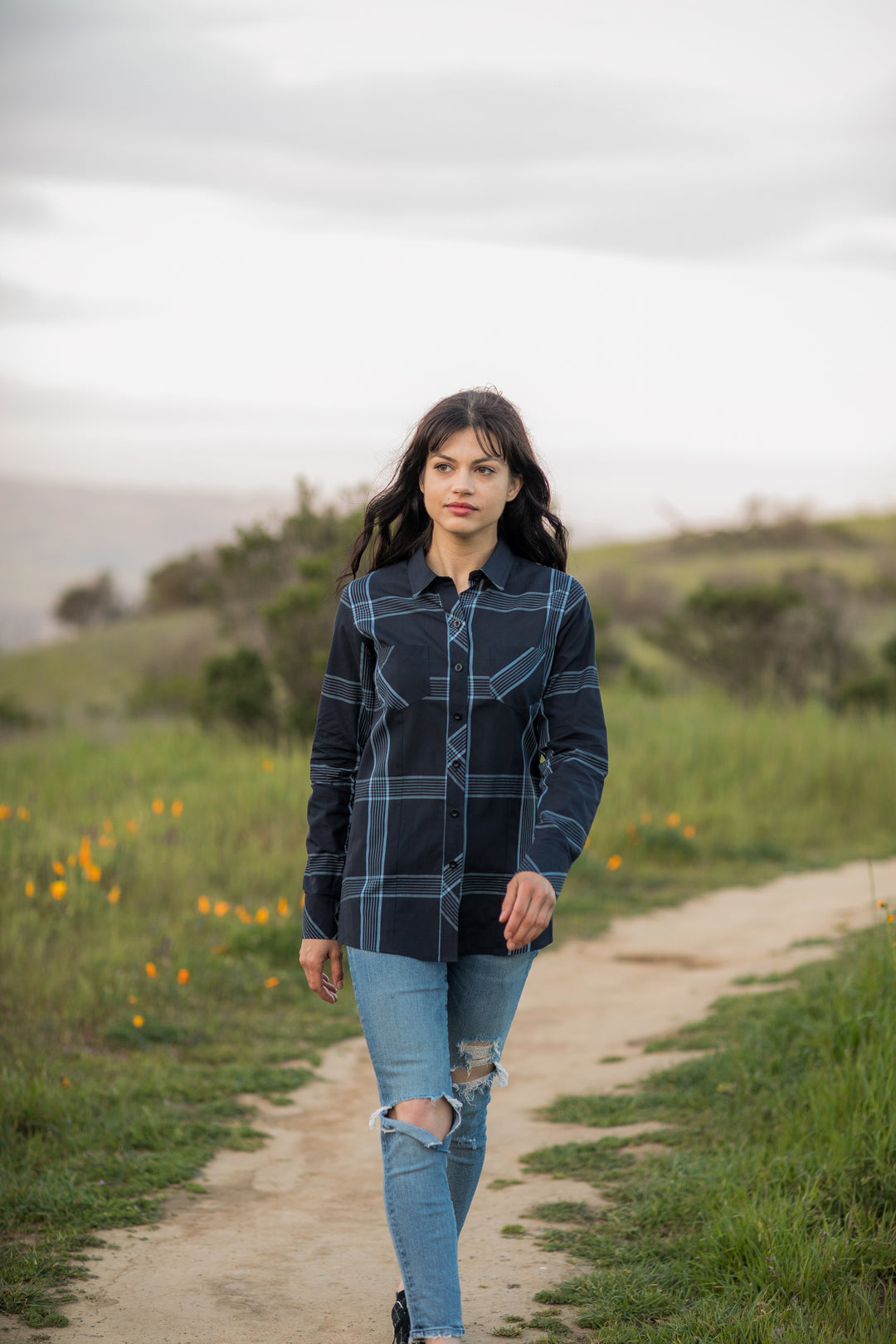 A woman with long dark hair walks along a dirt path in a grassy field with wildflowers, wearing the Women&