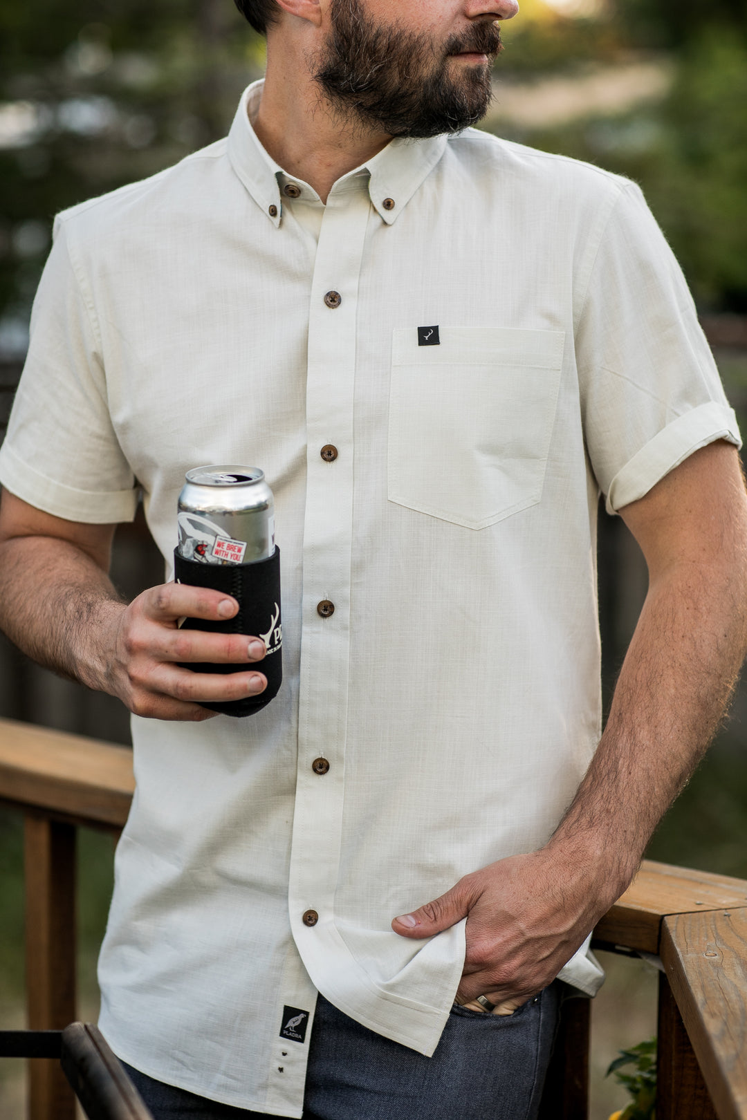 A man stands outdoors by a wooden railing, holding a canned drink with a black koozie and wearing the Men&