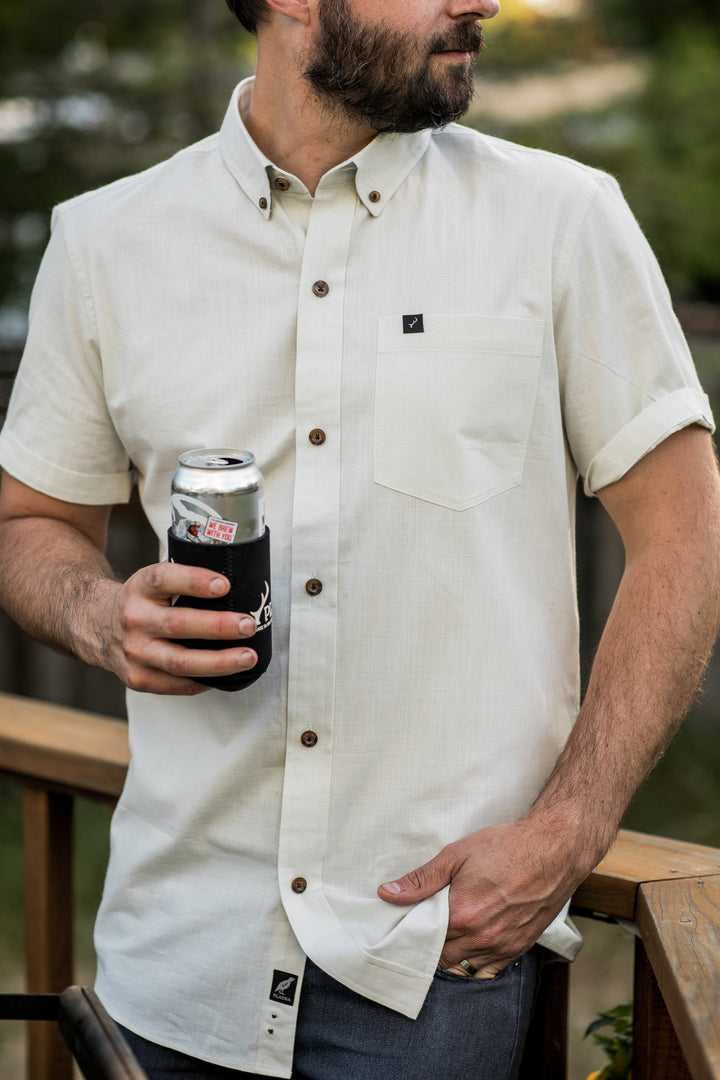 A man stands outdoors by a wooden railing, holding a canned drink with a black koozie and wearing the Men&