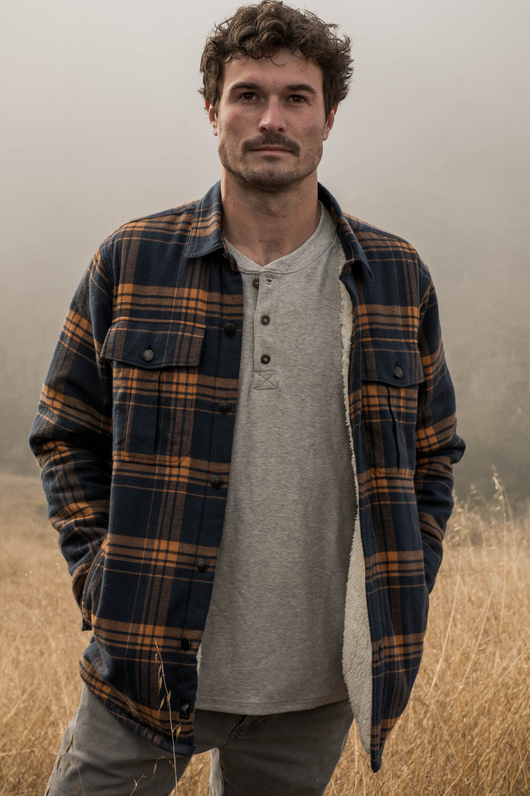 A man with curly hair and a mustache stands in a field of dry grass, wearing a gray shirt and the Men&