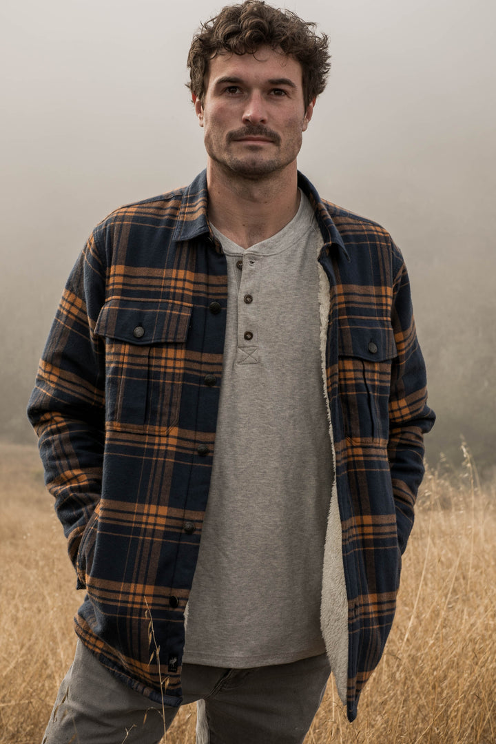 A man with curly hair and a mustache stands in a field of dry grass, wearing a gray shirt and the Men&