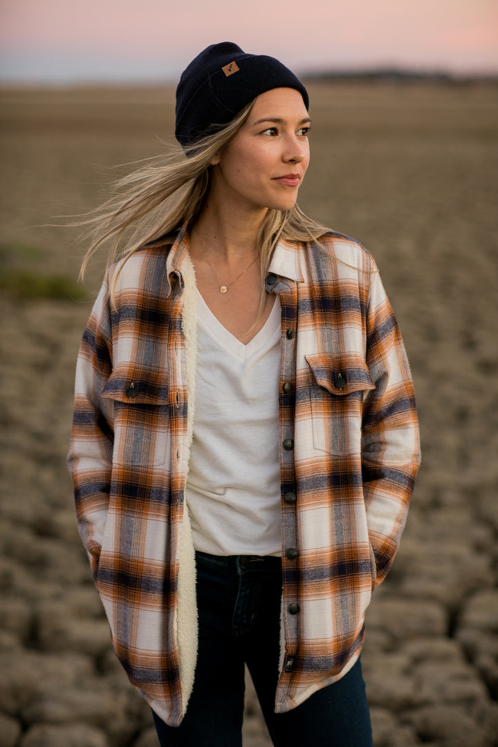 Woman in Pladra’s Northwood Sherpa flannel jacket, Peak White, and beanie stands on cracked earth at sunset.