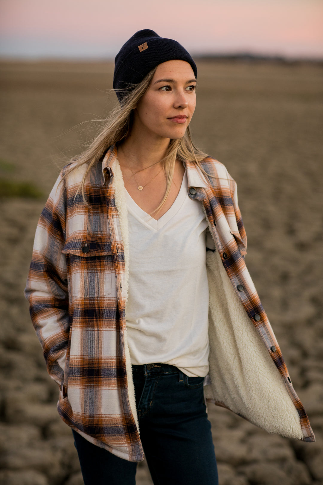 A woman in a Pladra Northwood Sherpa Insulated Flannel Jacket—Peak White—stands on cracked ground at sunset, gazing.