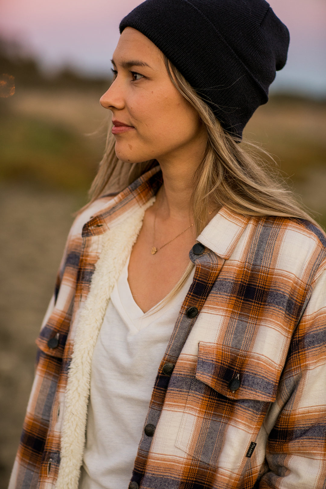 Woman in a black beanie and Pladra Northwood Sherpa Flannel Jacket, Peak White, stands outdoors in soft evening light.