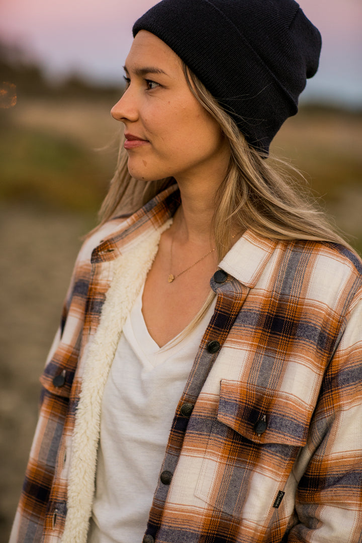 Woman in a black beanie and Pladra Northwood Sherpa Flannel Jacket, Peak White, stands outdoors in soft evening light.