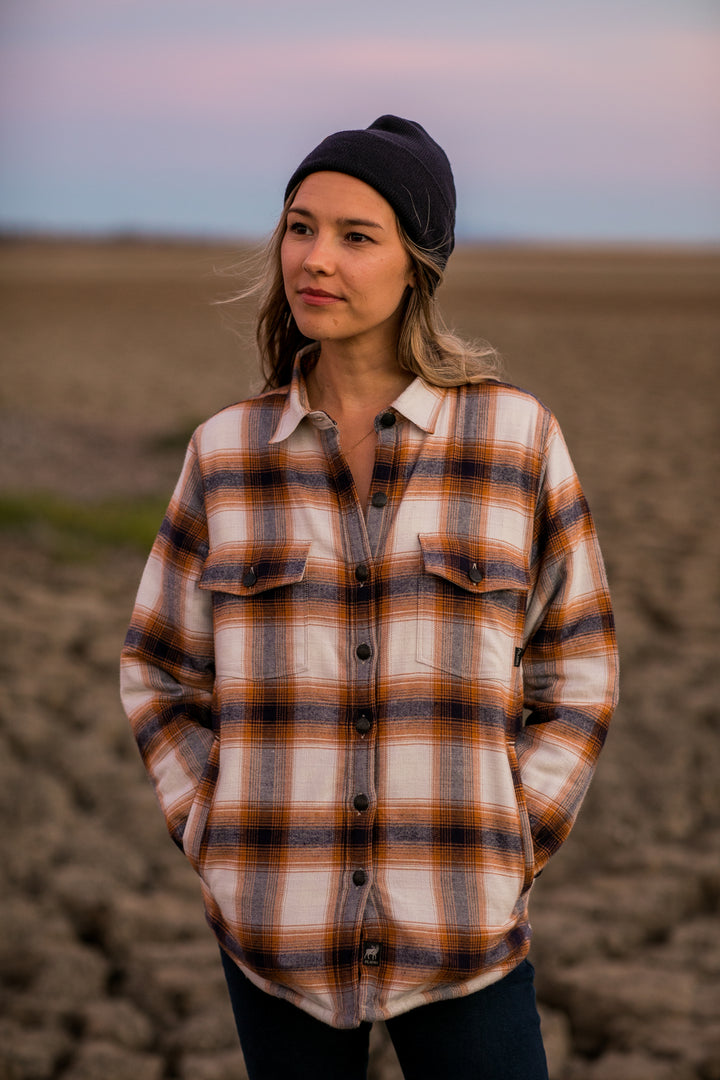 Woman in a Pladra Northwood Sherpa Flannel Jacket—Peak White—stands outdoors at sunset, hands in pockets.