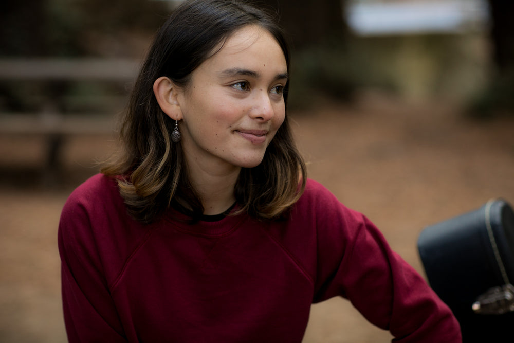 A young person with shoulder-length brown hair, wearing the Women's Crew Neck Sweatshirt in Pomegranate Red and dangling earrings, sits outside smiling gently, with a blurred backdrop of trees and a bench.