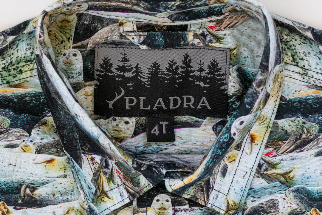 Close-up of the Toddler Cubs Outdoor Aloha Shirt- Seals collar with a black PLADRA label featuring trees and an antler. Made from 100% cotton, the multicolored print includes seals and subtle mountain lion motifs. Size tag reads 4T.