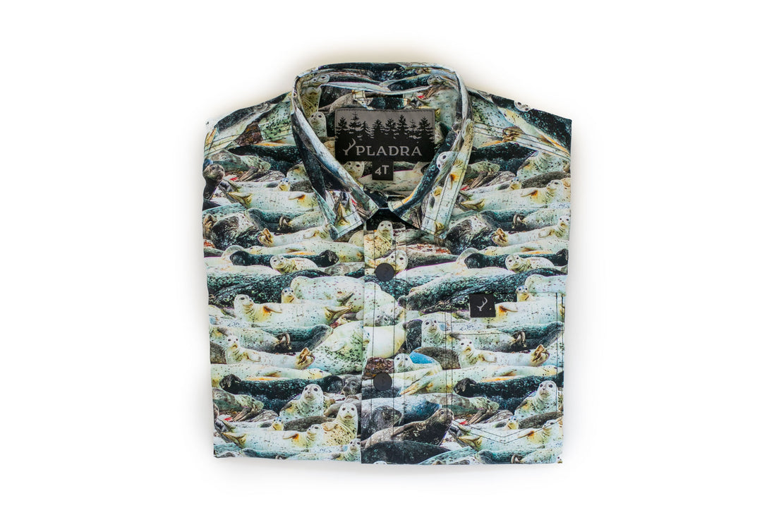 The Toddler Cubs Outdoor Aloha Shirt–Seals is a folded, 100% cotton button-up with a white background and an all-over print of realistic seals on rocks. The PLADRA label features a forest design.