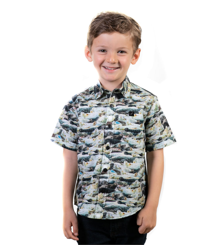 A young boy with short brown hair smiles while wearing the Toddler Cubs Outdoor Aloha Shirt- Seals, a 100% cotton, short-sleeved shirt with a vibrant seal pattern. He stands against a plain white background.
