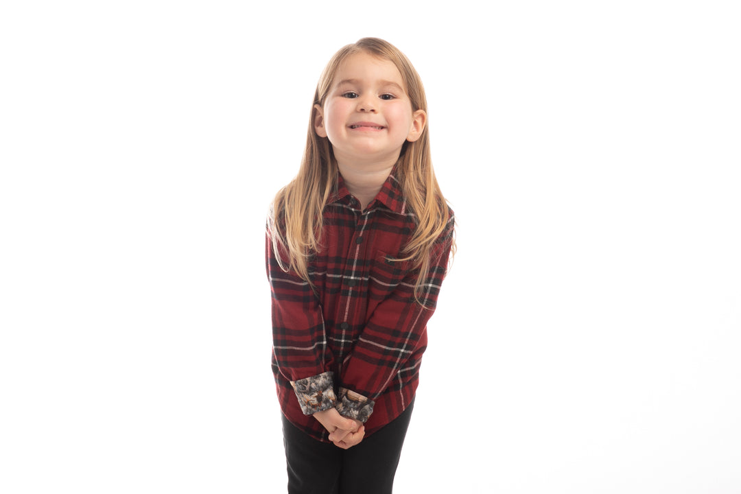 A young child with long blonde hair smiles and leans forward, wearing the Toddler Cubs Flannel Shirt—Rocky Red and black pants against a white background.