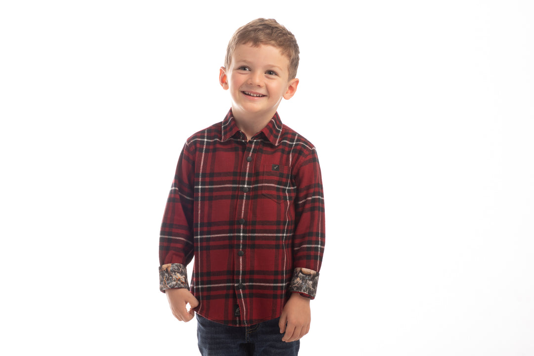 A young boy with short brown hair smiles against a plain white background, wearing the Toddler Cubs Flannel Shirt- Rocky Red, a premium cotton flannel in red and black plaid with rolled-up sleeves, paired with dark jeans.