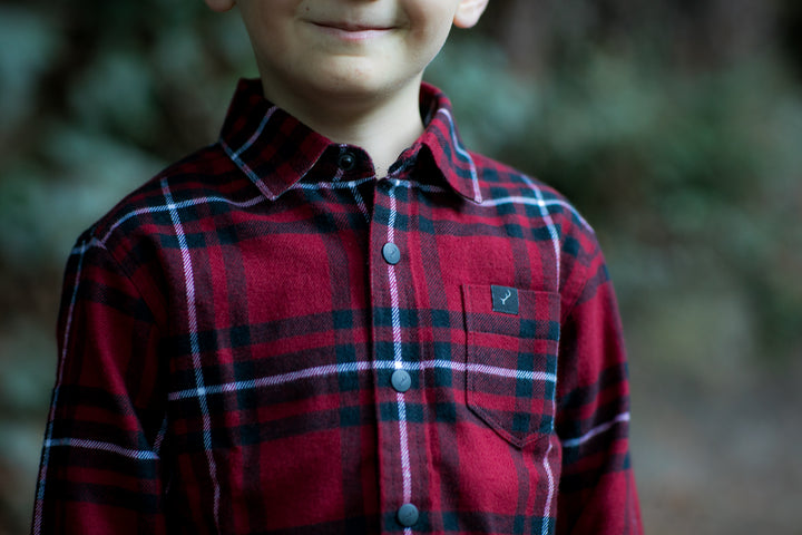 A child in the Toddler Cubs Flannel Shirt- Rocky Red smiles outdoors, with only the lower face and torso visible against a blurred natural background.