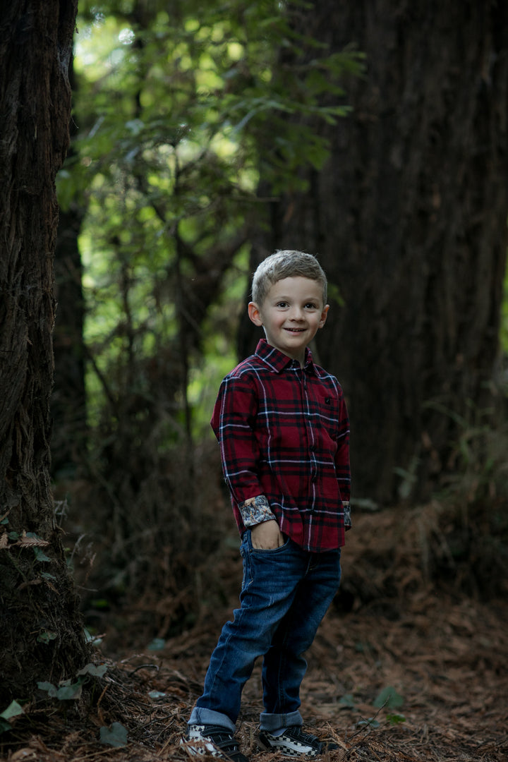 A young boy smiles outdoors in a forest, wearing the Toddler Cubs Flannel Shirt- Rocky Red and jeans. He stands among tall trees and green foliage with one hand in his pocket.