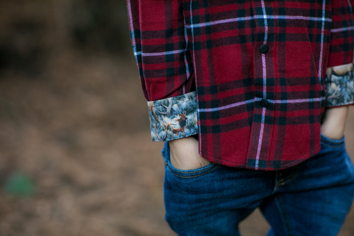 A toddler wears the Toddler Cubs Flannel Shirt- Rocky Red, featuring red and black plaid with rolled-up cuffs and blue jeans, standing outdoors with hands in pockets against a blurred background.