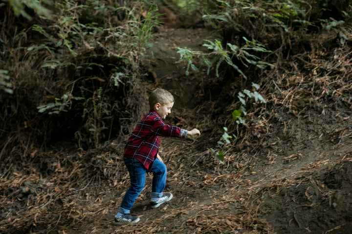 A young boy in a Toddler Cubs Flannel Shirt- Rocky Red, jeans, and sneakers climbs a dirt path through a forest surrounded by trees and leaves.