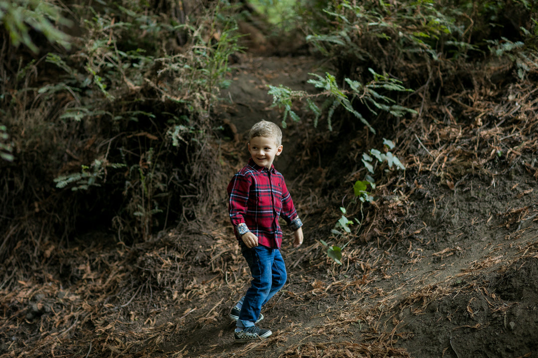 A young boy smiles as he walks along a forest path in the Toddler Cubs Flannel Shirt—Rocky Red, paired with jeans, surrounded by trees and fallen leaves.