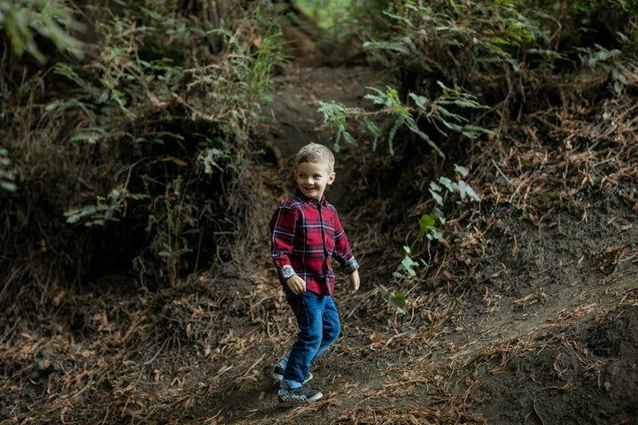A young boy smiles as he walks along a forest path in the Toddler Cubs Flannel Shirt—Rocky Red, paired with jeans, surrounded by trees and fallen leaves.