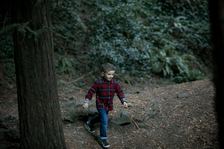A young boy wearing the Toddler Cubs Flannel Shirt- Rocky Red and jeans walks through a forested area with trees, rocks, and leafy ground cover.