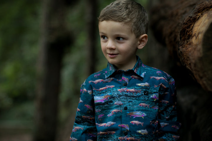 A toddler wearing the Pladra Toddler Cubs Printed Shirt - Trout stands outdoors near a large tree.