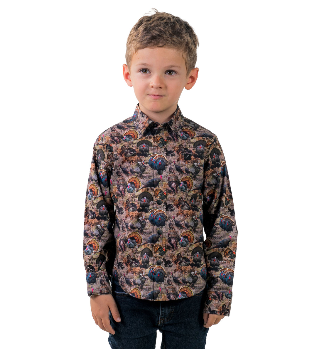 A young boy with short light brown hair wears the Toddler Cubs Printed Shirt- Turkeys, a long-sleeve featuring a colorful turkey and forest pattern. He stands facing forward with a neutral expression against a white background.