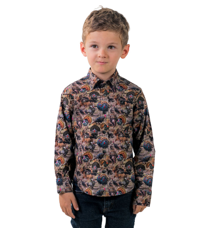 A young boy with short light brown hair wears the Toddler Cubs Printed Shirt- Turkeys, a long-sleeve featuring a colorful turkey and forest pattern. He stands facing forward with a neutral expression against a white background.