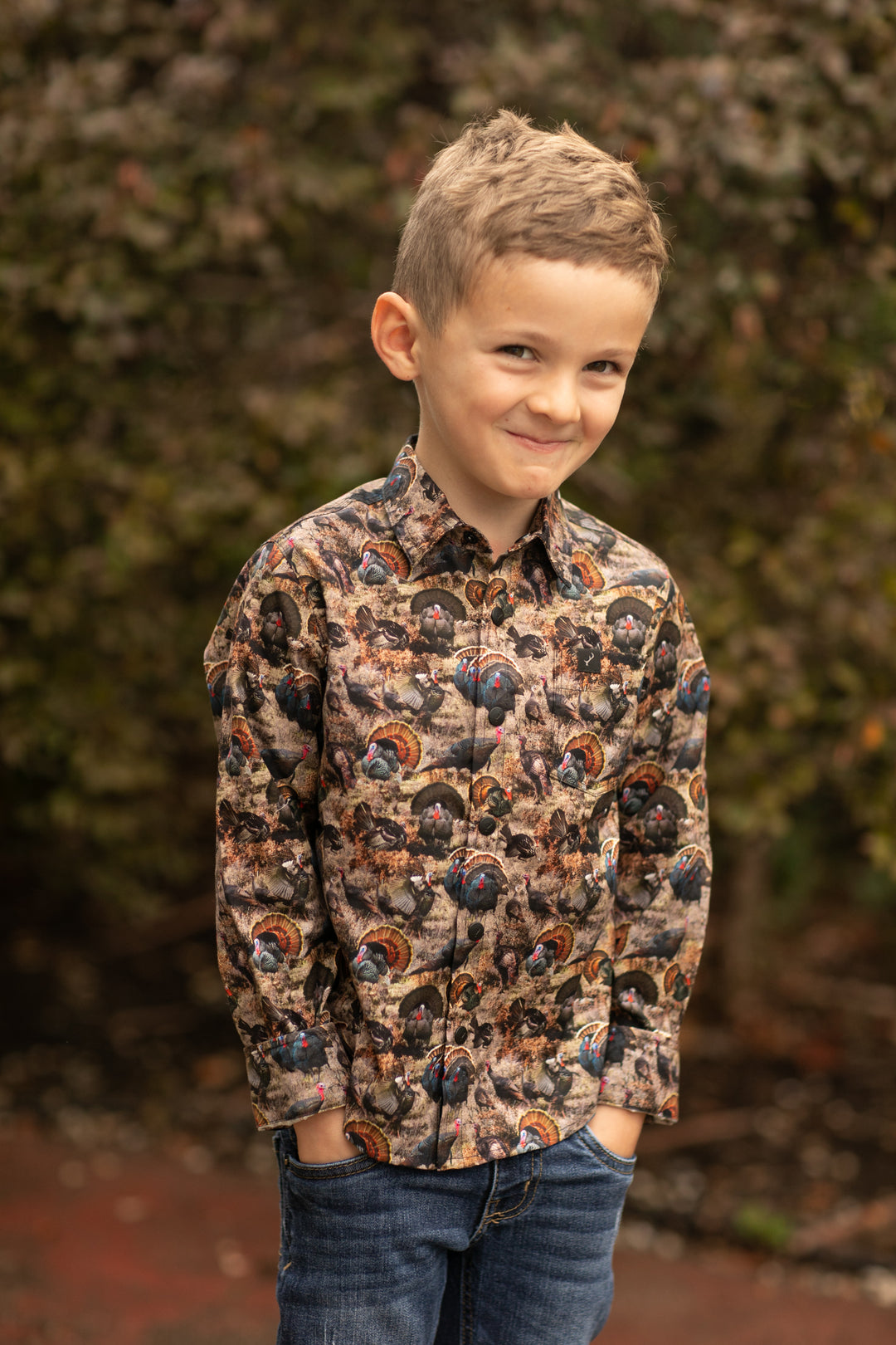 A young boy stands outdoors in front of leafy bushes, smiling slightly with his hands in his jeans pockets while wearing the Toddler Cubs Printed Shirt- Turkeys.