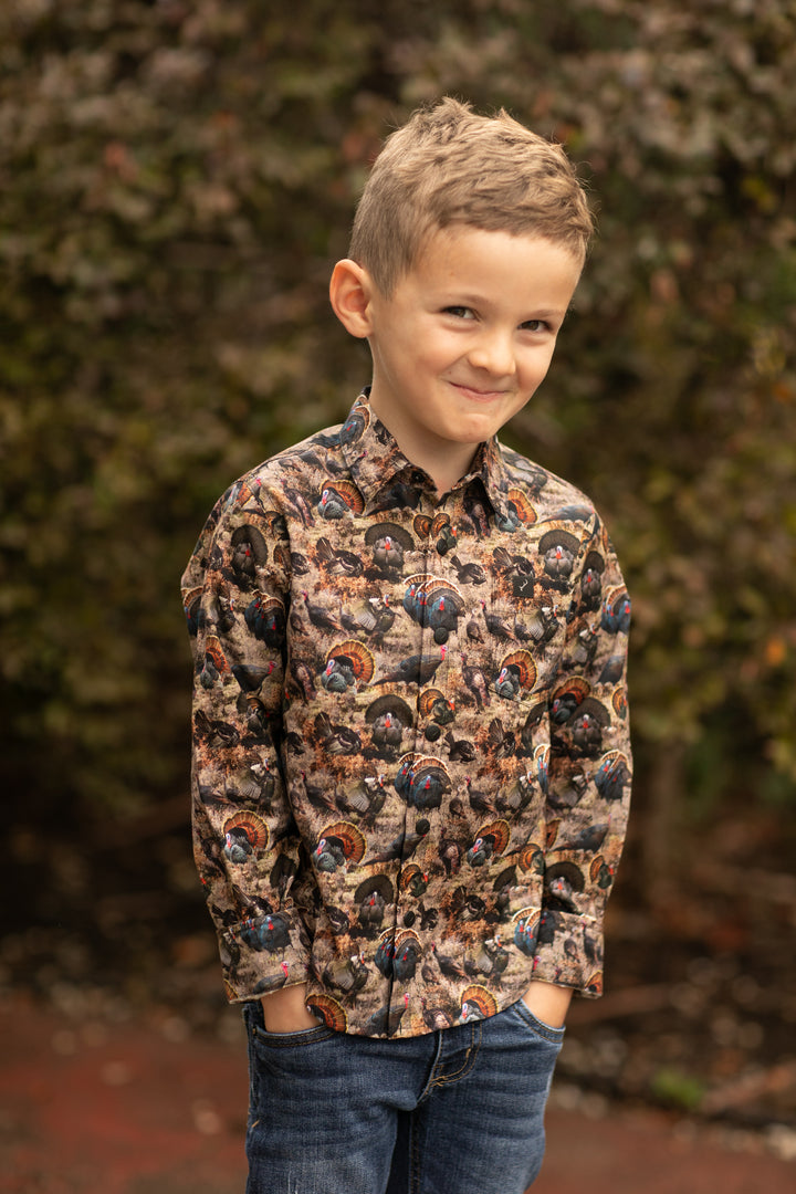 A young boy stands outdoors in front of leafy bushes, smiling slightly with his hands in his jeans pockets while wearing the Toddler Cubs Printed Shirt- Turkeys.