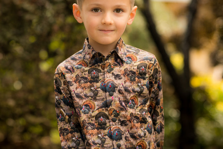 A young boy stands outdoors in the Toddler Cubs Printed Shirt- Turkeys, featuring a vibrant and detailed wild turkey print. The background shows blurred greenery and sunlight.