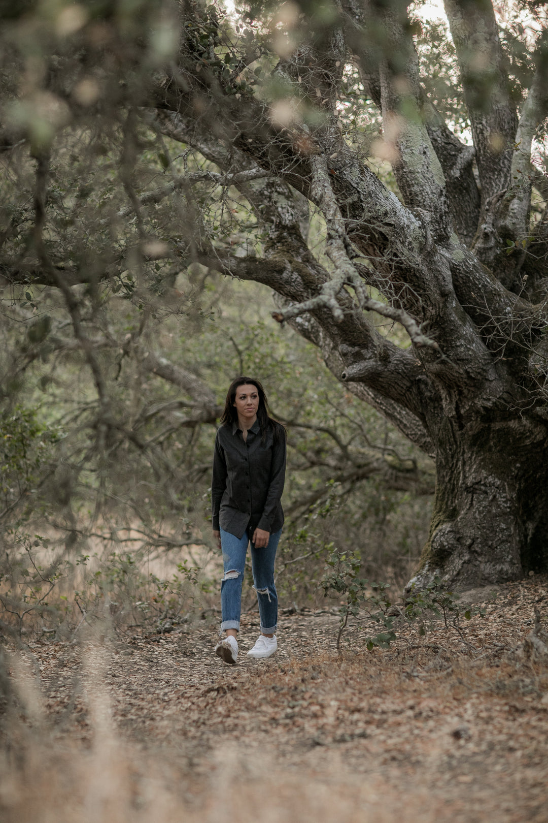 A woman in a black jacket, ripped jeans, and white shoes strolls alone through a forest; the Women’s Cascade Flannel Shirt - Jet Black Heather peeks from beneath her jacket, echoing the tones of the earthy, leaf-strewn ground.