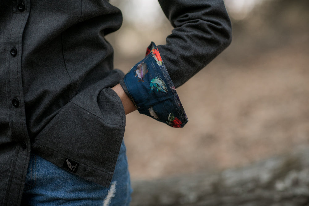 A woman stands outdoors in blue jeans and the Women's Cascade Flannel Shirt in Jet Black Heather, one hand in her pocket. The background is blurred and natural.
