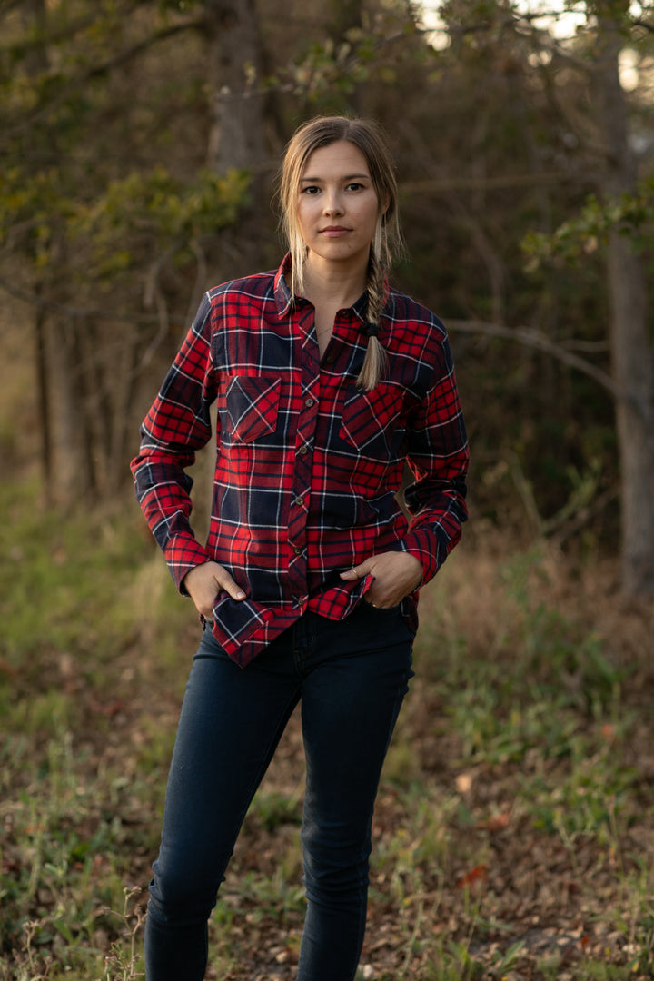A woman with blonde hair in two braids stands outdoors before trees, hands in her pockets, wearing the Women&