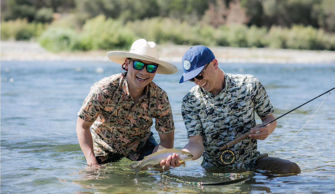 Two men kneel in shallow water fishing; one with a rod, the other holding a fish. Both wear Men&
